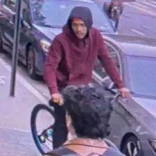 Security camera footage of one of the suspects, a male with light brown skin, is seen here wearing a maroon-colored, black-top hoodie and riding a bicycle.
