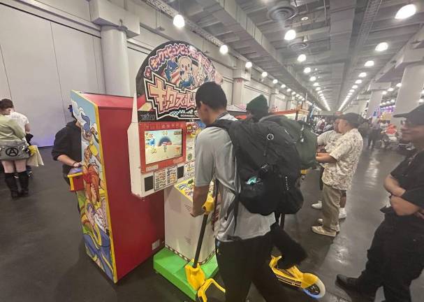 Convention goers line up and plat a Taito arcade racing game featuring colorful cartoon characters with some riders on yellow scooter controllers.